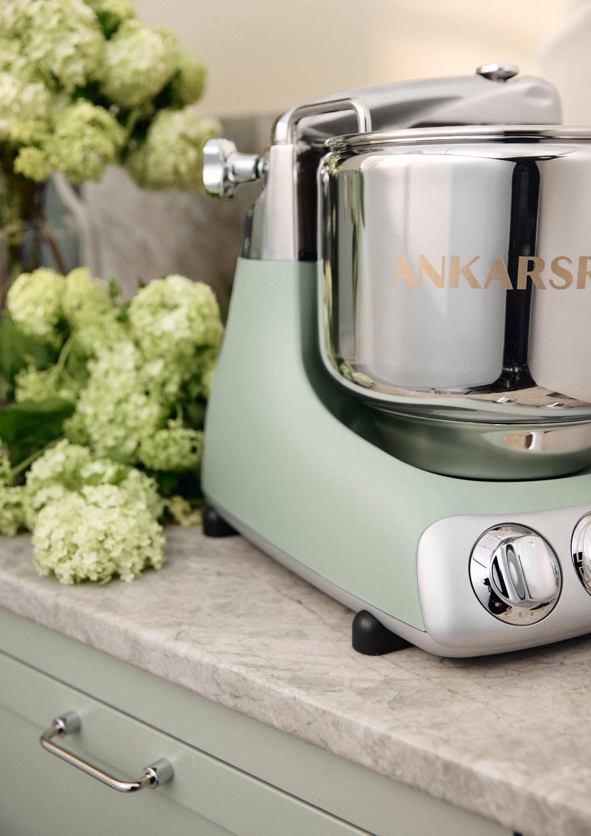 Nordic sage standing mixer with 'Ankarsrum' branding on a countertop with flowers in the background