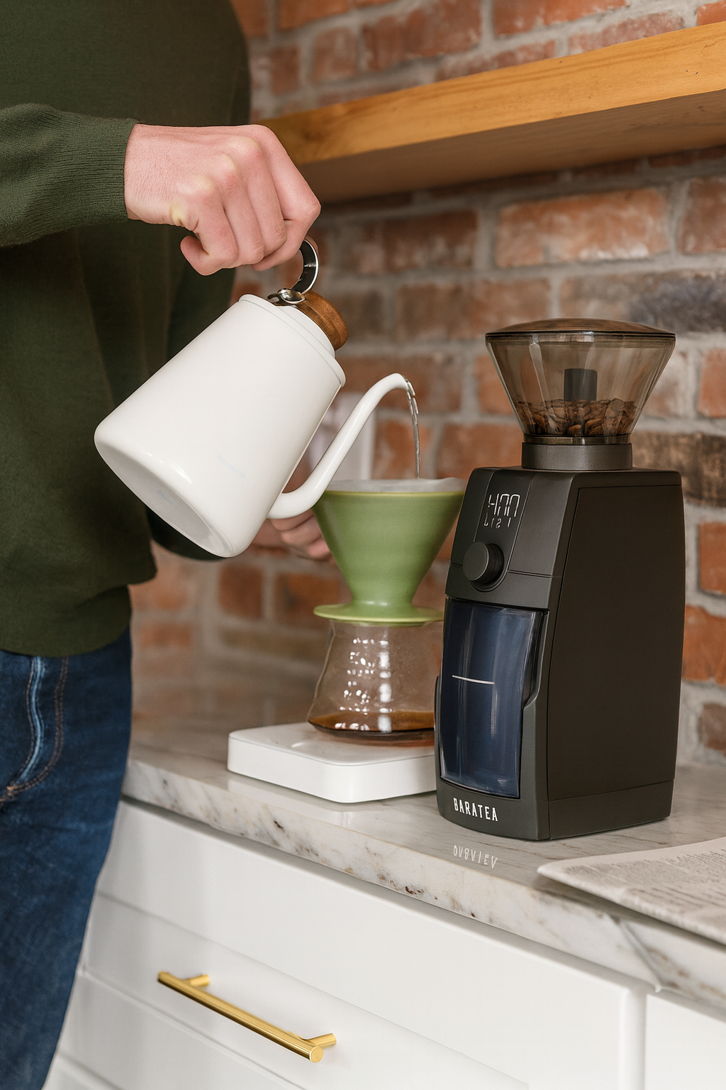 Person pouring coffee from a white kettle into a green coffee filter with a black coffee grinder on a kitchen counter.
