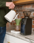 Person pouring coffee from a white kettle into a green coffee filter with a black coffee grinder on a kitchen counter.