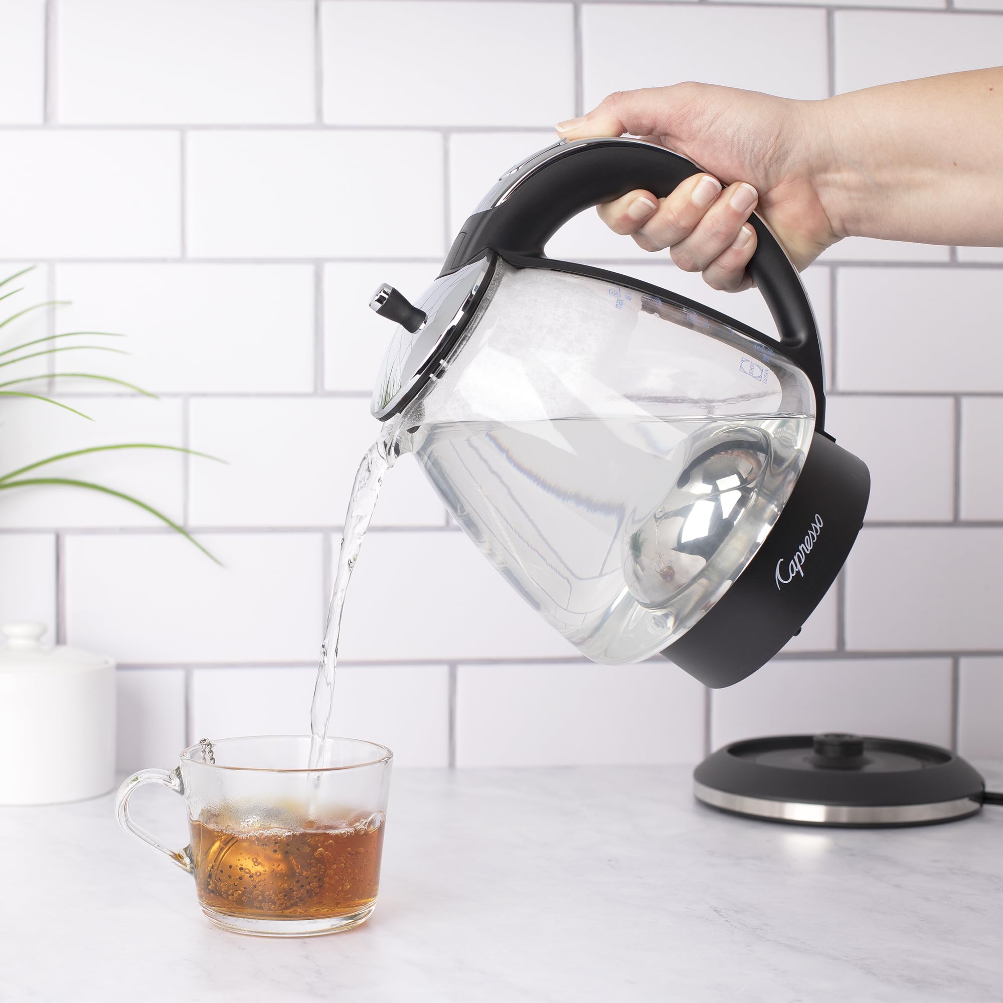 Person pouring water from a Black and polished chrome Capresso 48 oz electric kettle with black handle into a glass mug on a kitchen counter.