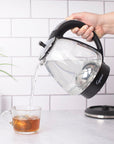 Person pouring water from a Black and polished chrome Capresso 48 oz electric kettle with black handle into a glass mug on a kitchen counter.