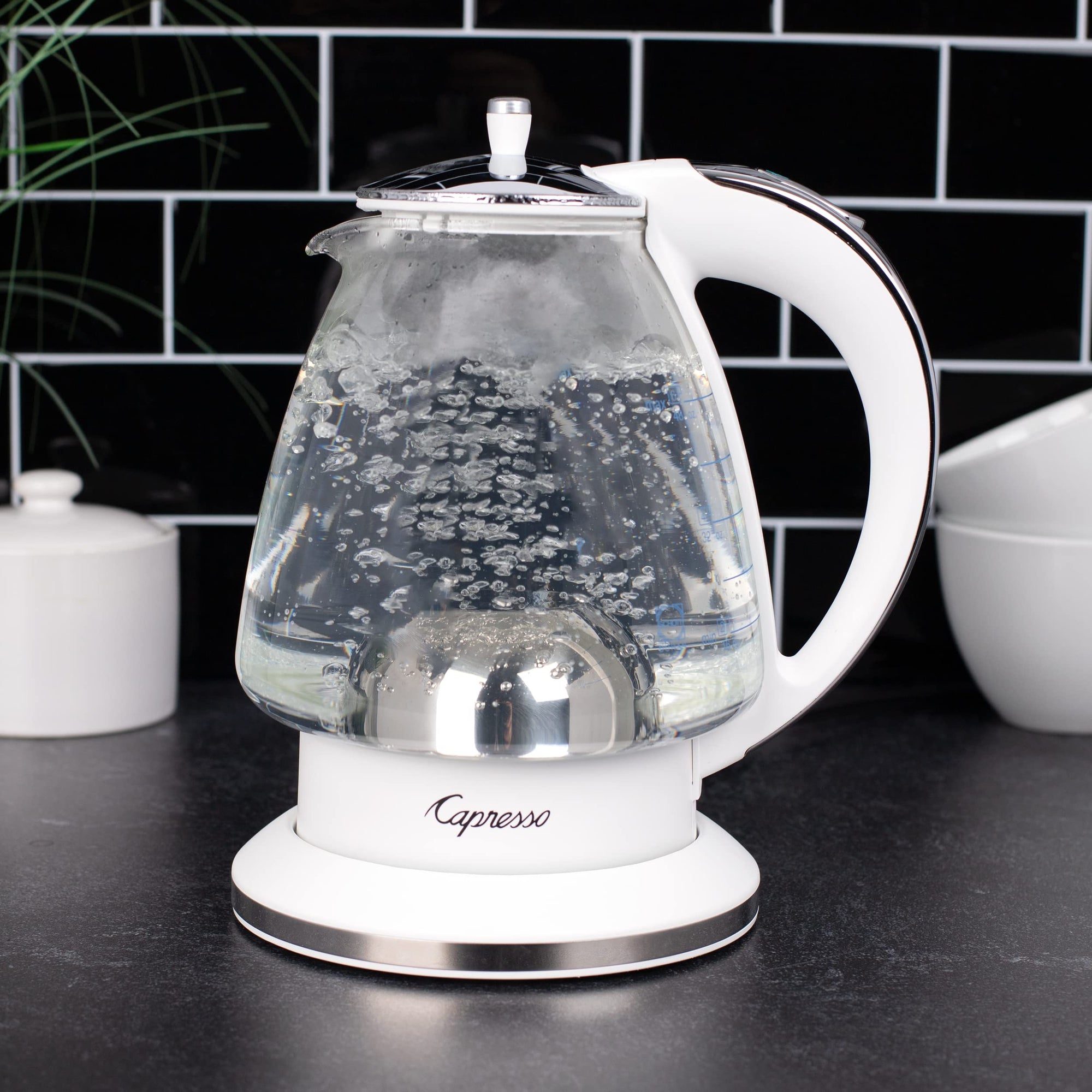 White and polished chrome Capresso 48 oz electric kettle with water boiling on a kitchen counter with a tiled wall background