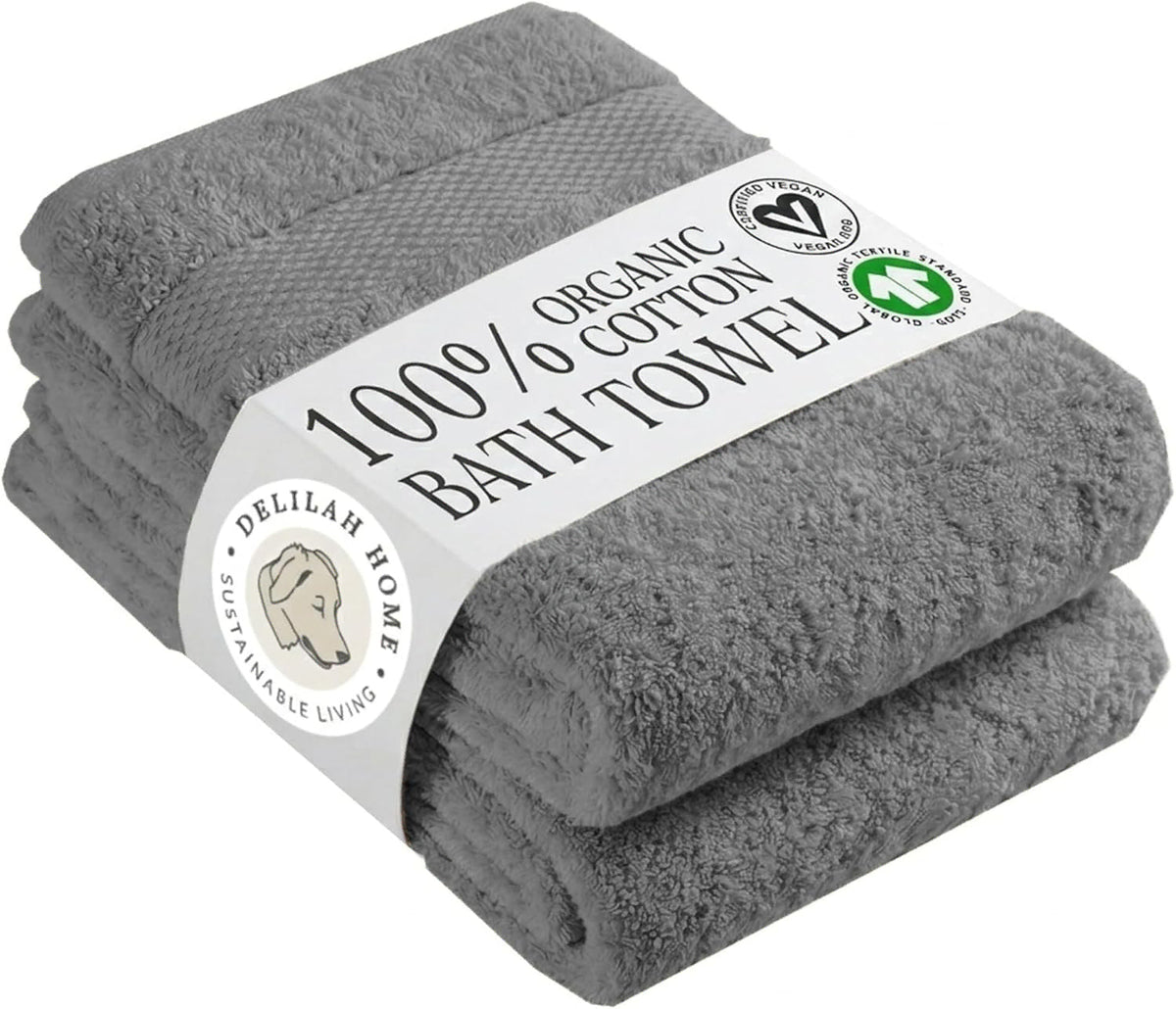 A stack of two folded gray cotton bath towels wrapped with a white paper label that reads '100% Organic Bath Towel' and shows the Delilah Home brand logo.