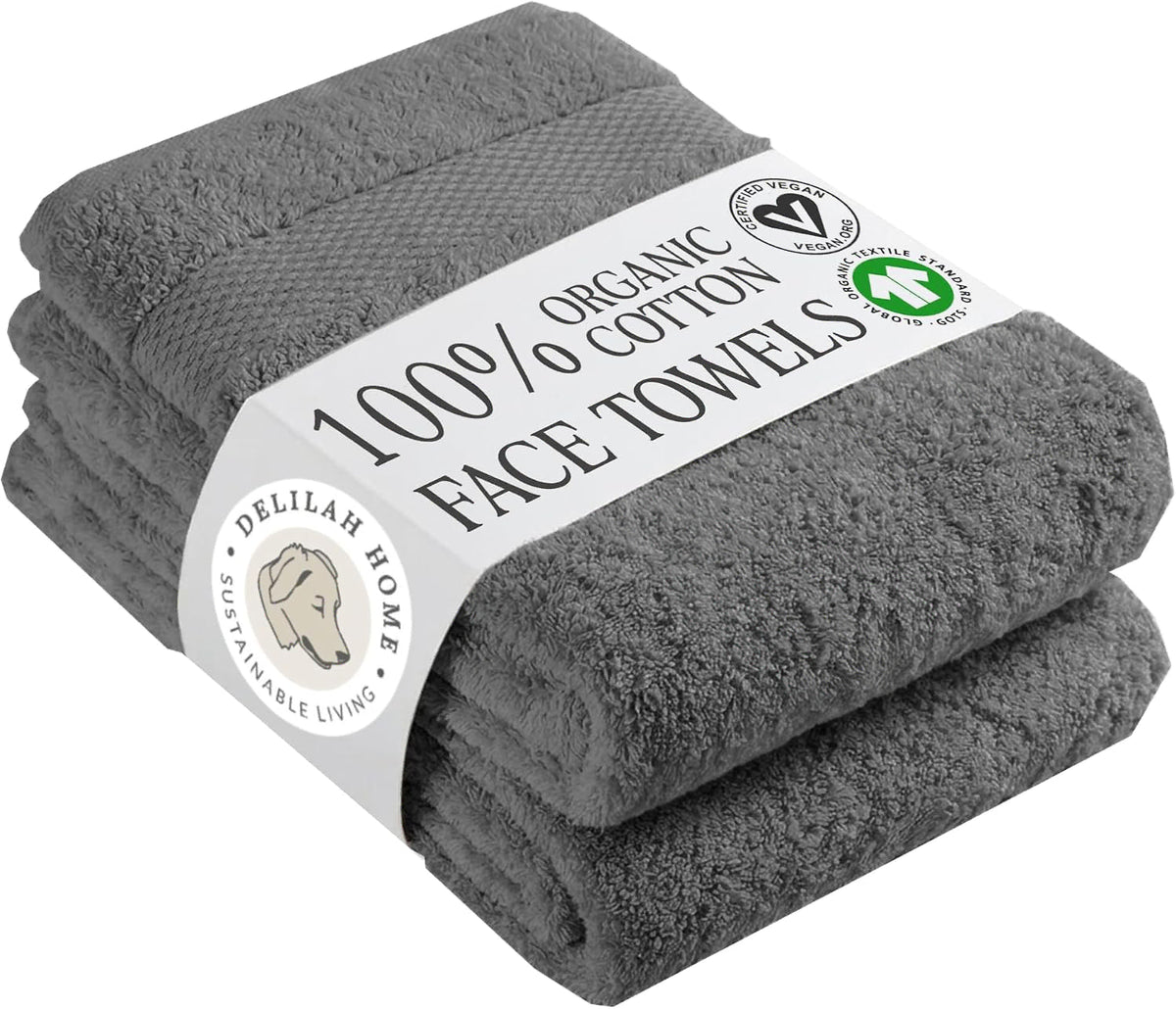 A stack of two folded gray cotton face towels wrapped in a white paper band that reads '100% Organic Cotton Face Towels' with certification logos and the Delilah Home brand name.