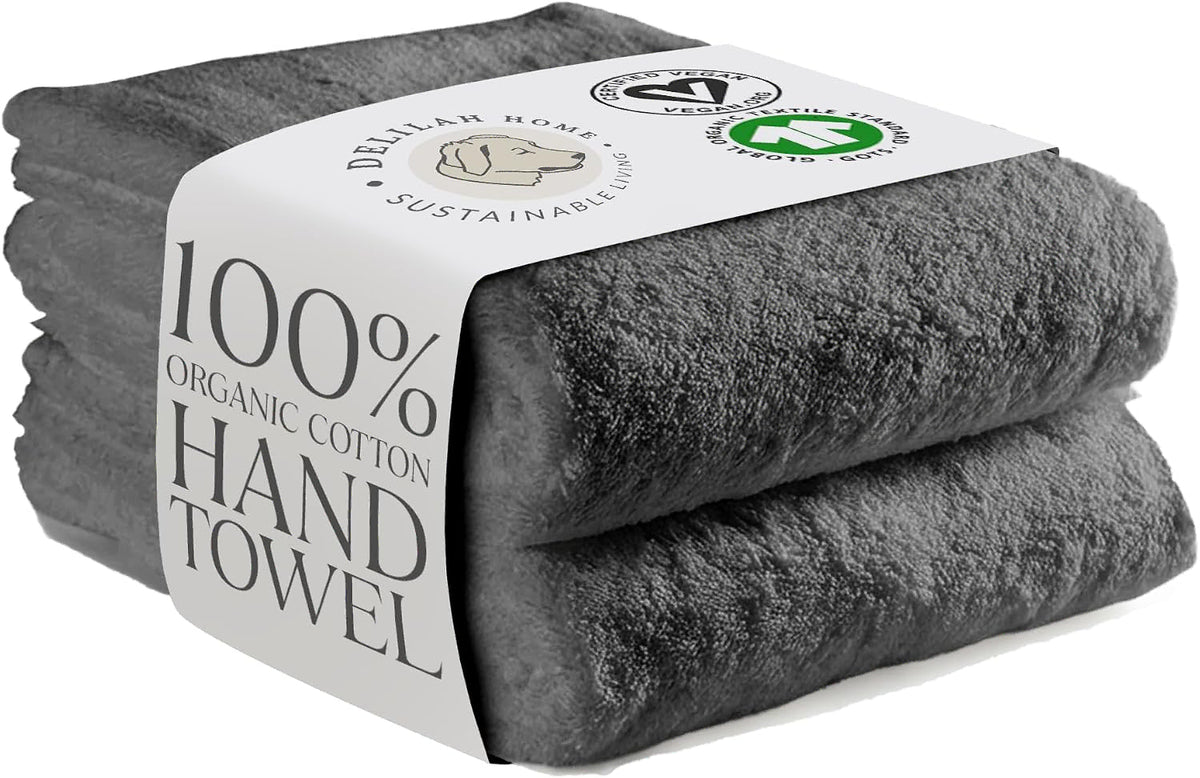 Two folded dark gray organic cotton hand towels wrapped in a white paper band showing certification logos and the text '100% ORGANIC COTTON HAND TOWEL'.