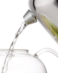 Clear glass carafe being filled with liquid from a white electric kettle on a white background