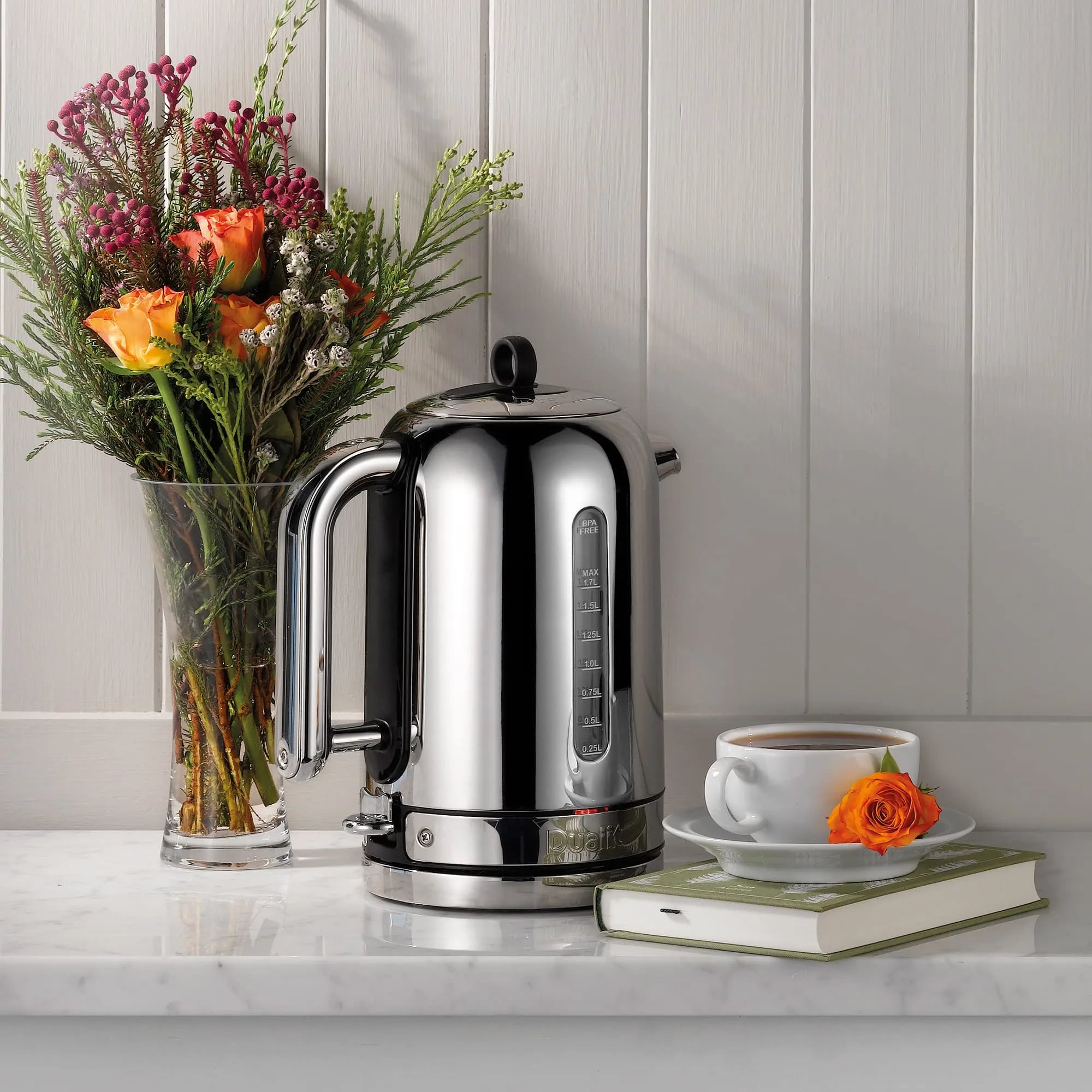 Silver kettle on a kitchen counter with flowers and a cup of coffee.