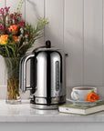 Silver kettle on a kitchen counter with flowers and a cup of coffee.