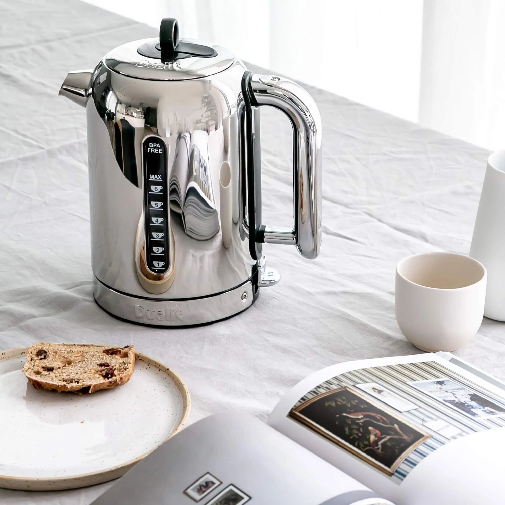 Stainless steel kettle on a white surface with a newspaper and bread