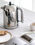 Stainless steel kettle on a white surface with a newspaper and bread
