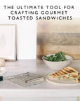 Toasted sandwich on a plate with a bowl of greens, bread, and a sandwich cage on a kitchen counter.