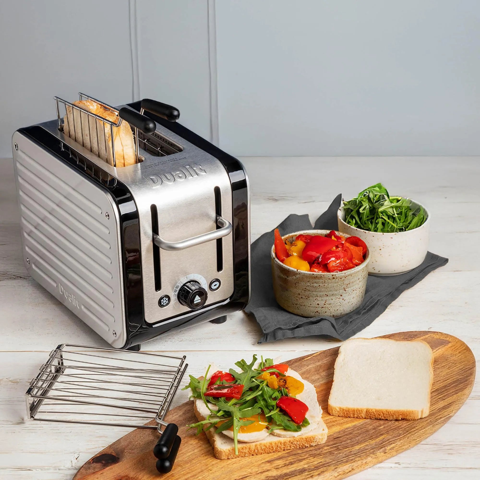 A silver and black Dualit 2-slice toaster sits on a white wooden table, with bread slots open and toast emerging. Surrounding the toaster are bowls of salad, sliced white bread, and a wooden board, all on a light tabletop.