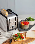 A silver and black Dualit 2-slice toaster sits on a white wooden table, with bread slots open and toast emerging. Surrounding the toaster are bowls of salad, sliced white bread, and a wooden board, all on a light tabletop.