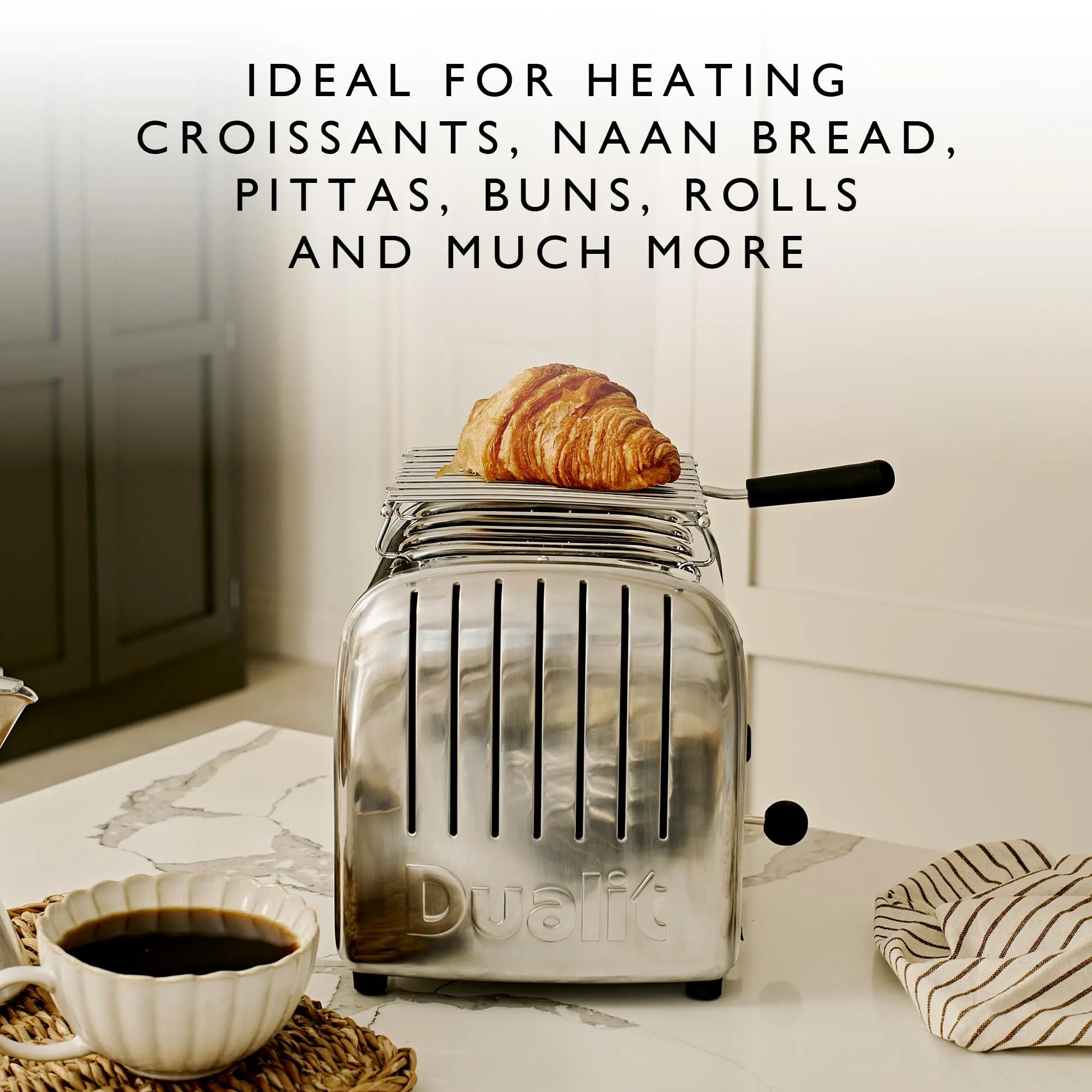 Silver toaster with a croissant on top of warming rack, placed on a kitchen counter with a cup of coffee.