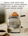 Silver toaster with a croissant on top of warming rack, placed on a kitchen counter with a cup of coffee.