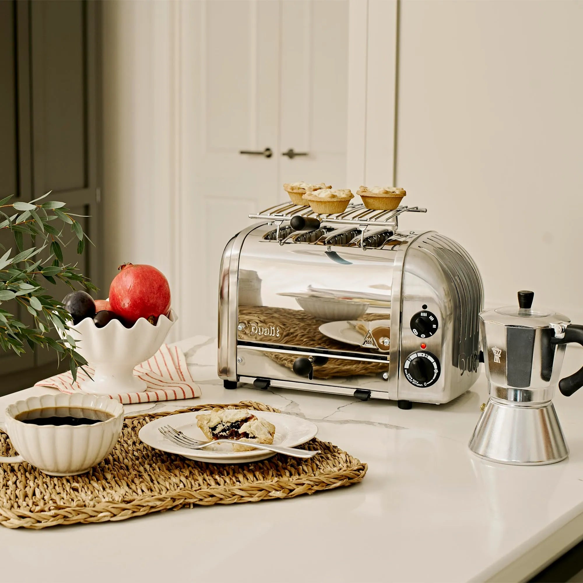 Silver toaster with a warming rack on a kitchen counter with a plate of food, coffee, and a plant.