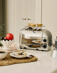 Silver toaster with a warming rack on a kitchen counter with a plate of food, coffee, and a plant.