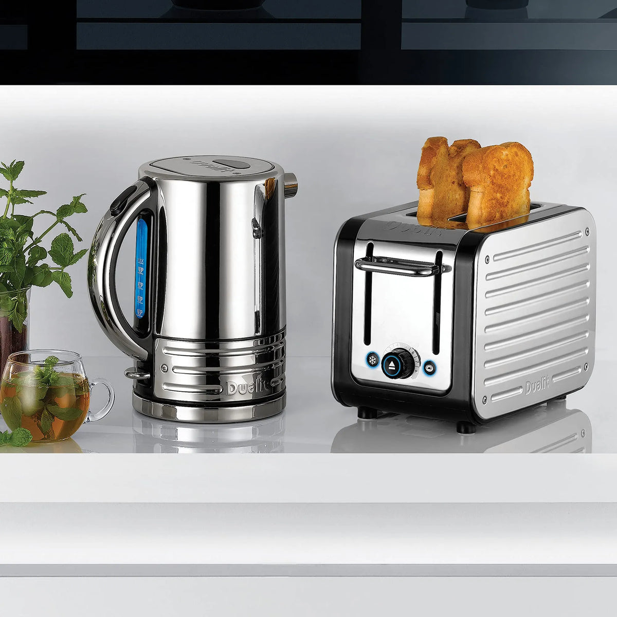 A Dualit stainless steel electric kettle and a matching toaster with two slices of toast, displayed on a white countertop with a small plant and glass of tea in the background.