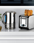 A Dualit stainless steel electric kettle and a matching toaster with two slices of toast, displayed on a white countertop with a small plant and glass of tea in the background.