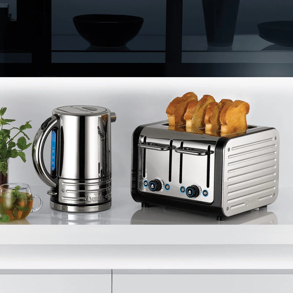 A stainless steel electric kettle and a matching two-slot toaster with four slices of toast, set on a white countertop in a modern kitchen scene.