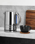 Silver electric kettle on a kitchen counter with a black mug and wooden cutting board.