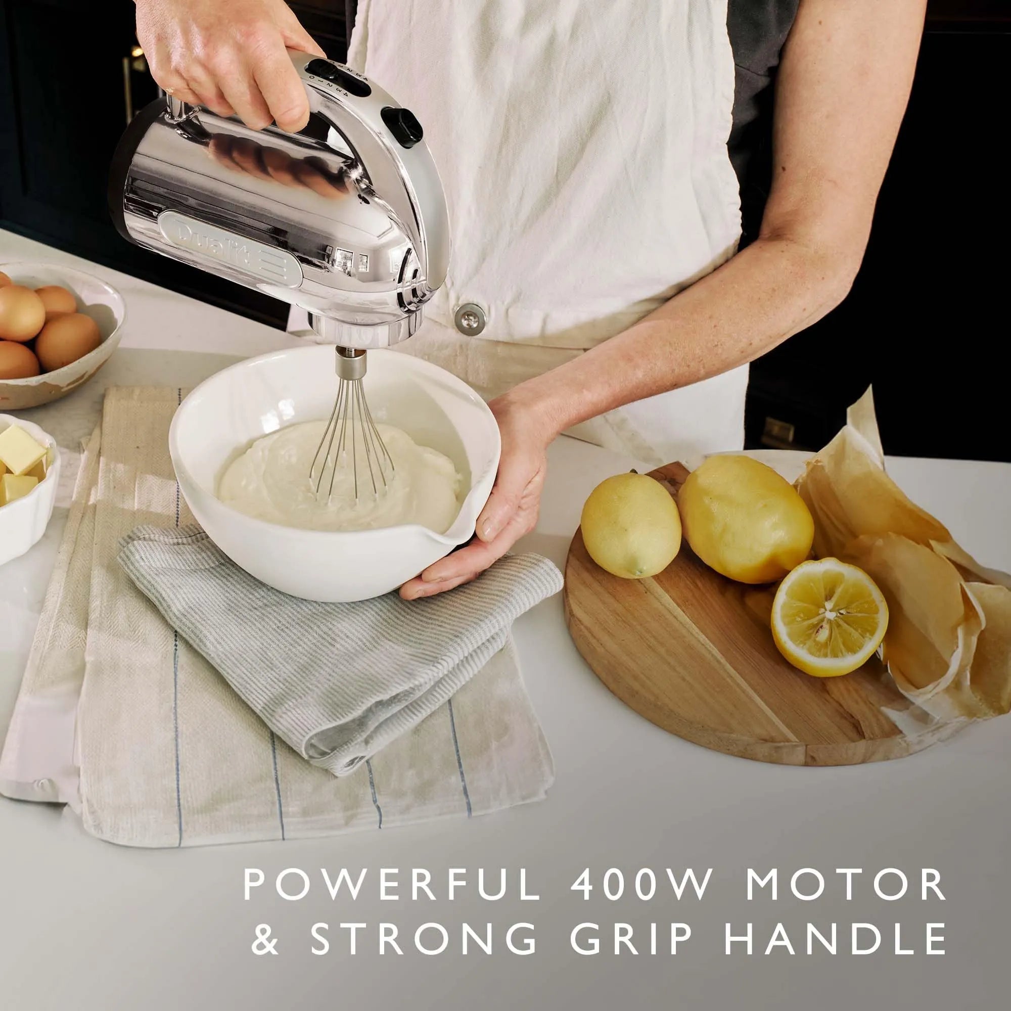 A person in a white apron uses a silver and black countertop hand mixer to whip cream in a white bowl. The scene includes eggs, butter, lemons, and parchment paper on a kitchen counter, with text at the bottom reading 'POWERFUL 400W MOTOR &amp; STRONG GRIP HANDLE'.