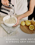 A person in a white apron uses a silver and black countertop hand mixer to whip cream in a white bowl. The scene includes eggs, butter, lemons, and parchment paper on a kitchen counter, with text at the bottom reading 'POWERFUL 400W MOTOR & STRONG GRIP HANDLE'.