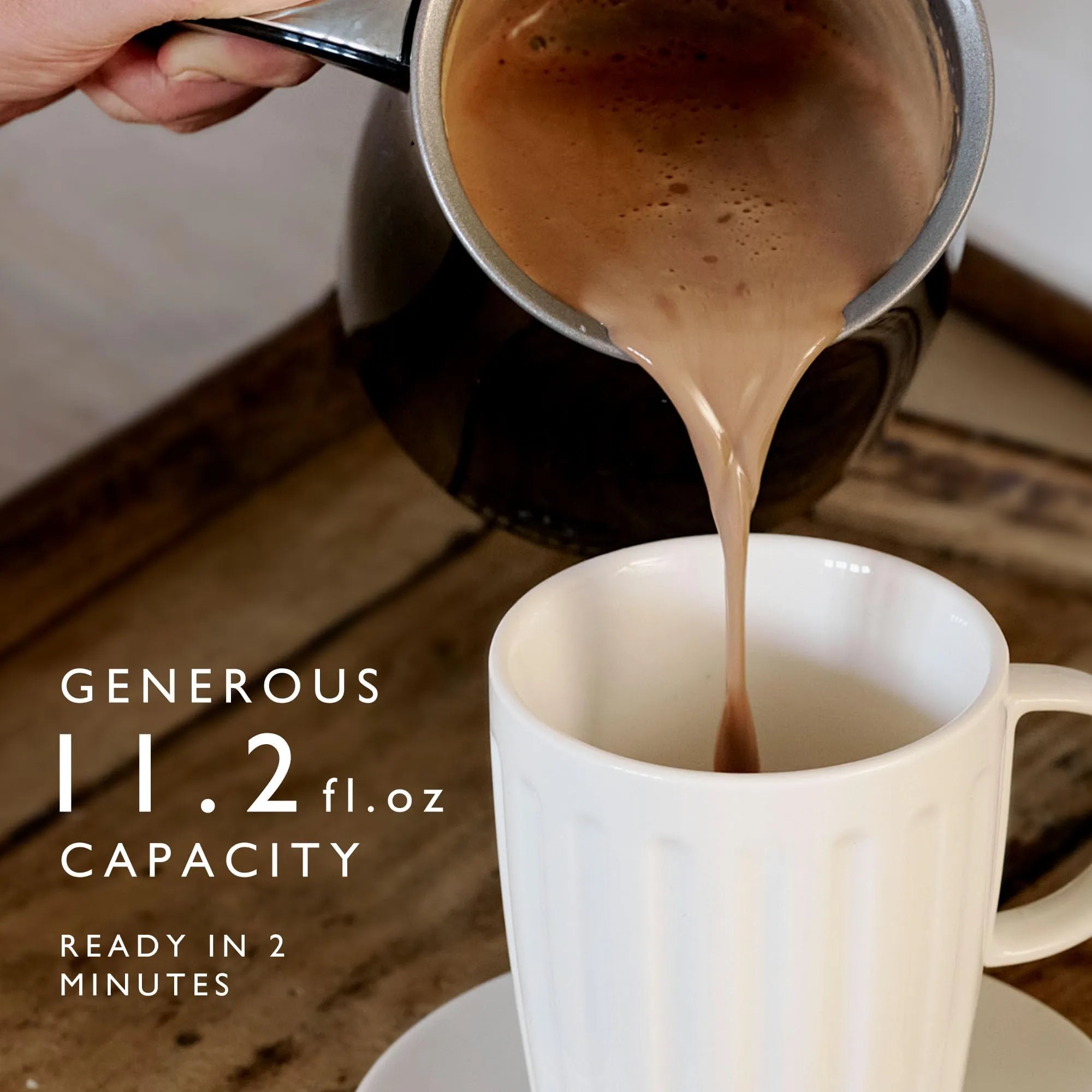 A close-up of a hand pouring hot coffee from a black milk frother into a white ribbed ceramic mug on a wooden table. White text on the left reads 'GENEROUS 11.2 fl oz capacity.