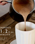 A close-up of a hand pouring hot coffee from a black milk frother into a white ribbed ceramic mug on a wooden table. White text on the left reads 'GENEROUS 11.2 fl oz capacity.