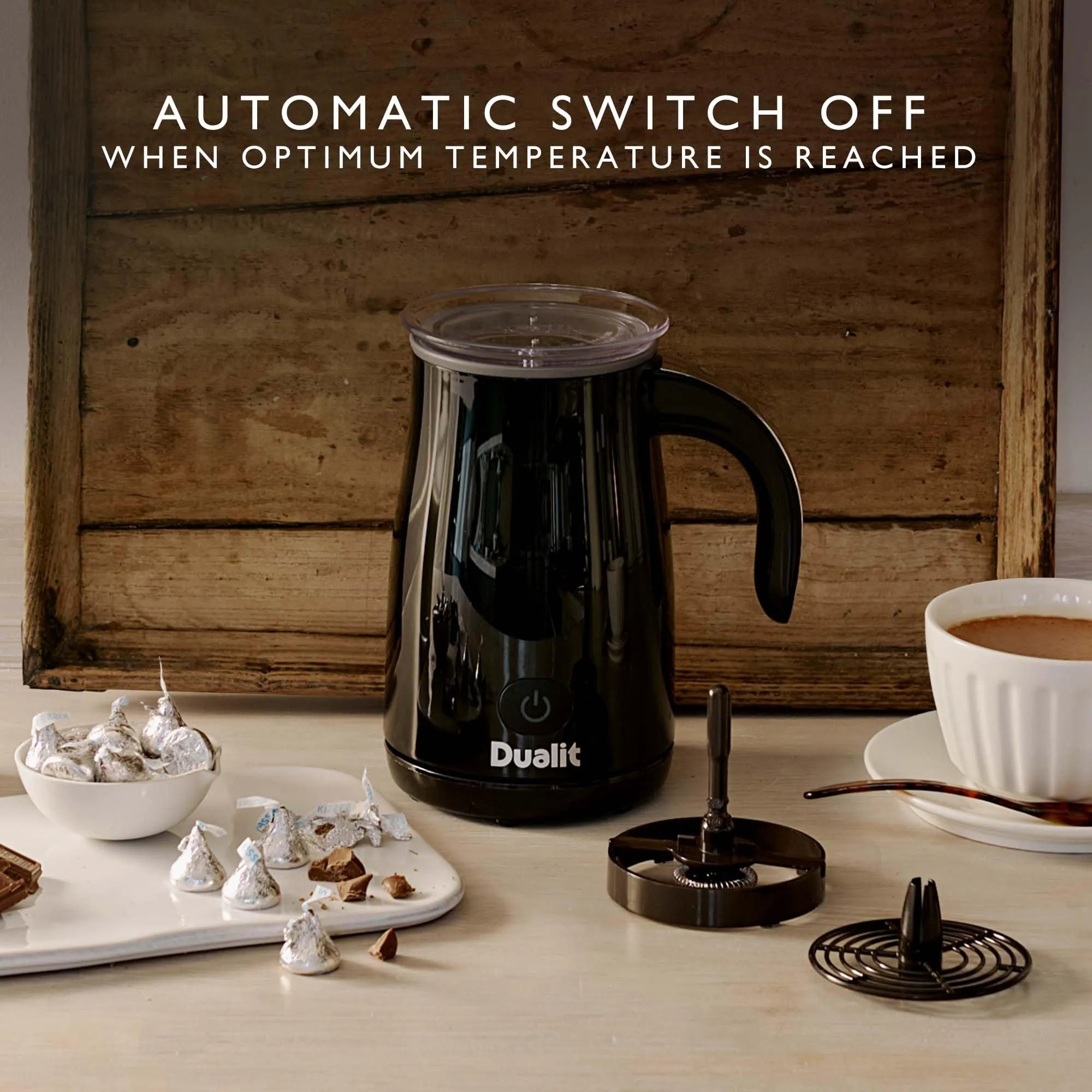 A black Dualit automatic milk frother with a clear lid sits on a tabletop in a styled kitchen scene, with its metal filter components placed beside it and a cup of coffee and chocolate kisses in the background. Text at the top reads: 'AUTOMATIC SWITCH OFF WHEN OPTIMUM TEMPERATURE IS REACHED'.