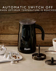 A black Dualit automatic milk frother with a clear lid sits on a tabletop in a styled kitchen scene, with its metal filter components placed beside it and a cup of coffee and chocolate kisses in the background. Text at the top reads: 'AUTOMATIC SWITCH OFF WHEN OPTIMUM TEMPERATURE IS REACHED'.
