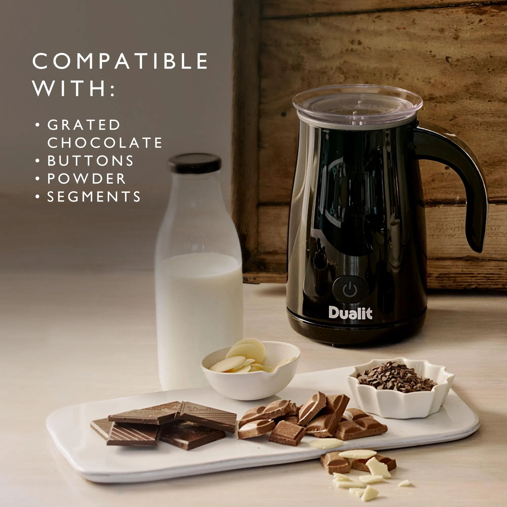 A Dualit milk frother in black with a clear lid and black handle, shown on a tabletop next to a glass bottle of milk, and chocolate pieces. Text on the left reads 'COMPATIBLE WITH: GRATED CHOCOLATE, BUTTONS, POWDER, SEGMENTS'.