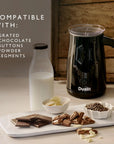 A Dualit milk frother in black with a clear lid and black handle, shown on a tabletop next to a glass bottle of milk, and chocolate pieces. Text on the left reads 'COMPATIBLE WITH: GRATED CHOCOLATE, BUTTONS, POWDER, SEGMENTS'.