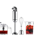 A stainless steel hand blender set with a tall immersion blender, a whisk attachment, a clear blending cup containing red liquid, and a clear measuring cup filled with strawberries, blackberries, and redcurrants on a white background.