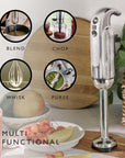 A silver and black immersion blender stands on a wooden board on a kitchen counter, with four circular inset images showing different blending functions: blend, chop, whisk, and puree. The scene includes fruits, a pink drink, and a potted plant in a kitchen setting.