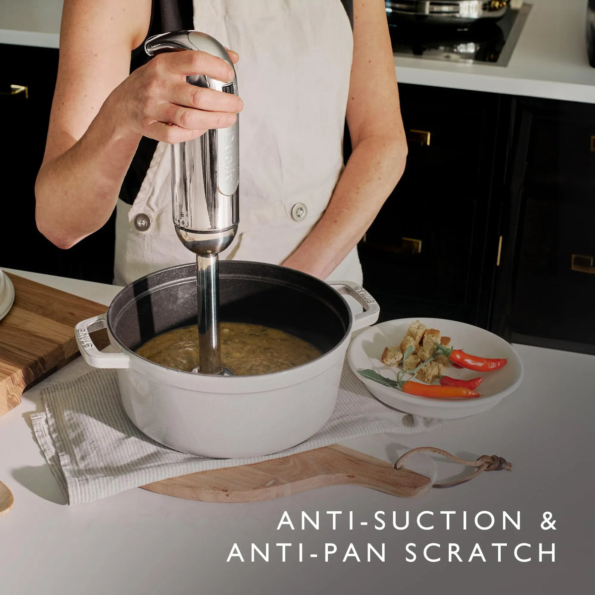 A person in a white apron uses an immersion blender to mix a soup in a white enameled cast iron pot on a kitchen counter. A plate of vegetables and the text 'ANTI-SUCTION &amp; ANTI-PAN SCRATCH' are visible.