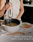 A person in a white apron uses an immersion blender to mix a soup in a white enameled cast iron pot on a kitchen counter. A plate of vegetables and the text 'ANTI-SUCTION & ANTI-PAN SCRATCH' are visible.