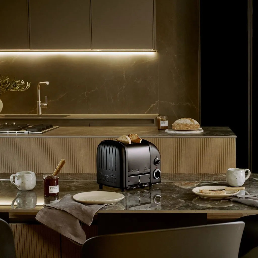 A black Dualit toaster sits on a polished kitchen countertop, surrounded by breakfast items like bread, a cup of coffee, and a small jar of jam in a modern, dimly lit kitchen setting.