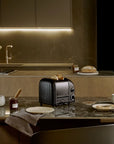 A black Dualit toaster sits on a polished kitchen countertop, surrounded by breakfast items like bread, a cup of coffee, and a small jar of jam in a modern, dimly lit kitchen setting.
