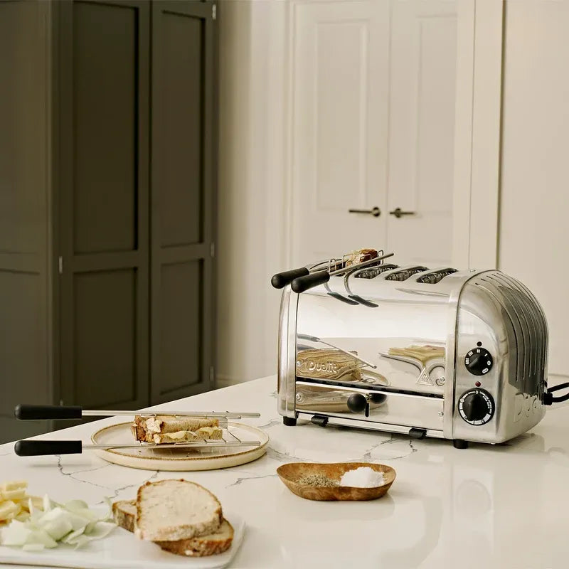 A stainless steel 4-slice toaster sits on a white marble kitchen countertop, with bread slices, onions, and a small dish of salt arranged nearby. The background shows a cream-colored paneled door and dark gray cabinets.