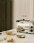 A stainless steel 4-slice toaster sits on a white marble kitchen countertop, with bread slices, onions, and a small dish of salt arranged nearby. The background shows a cream-colored paneled door and dark gray cabinets.