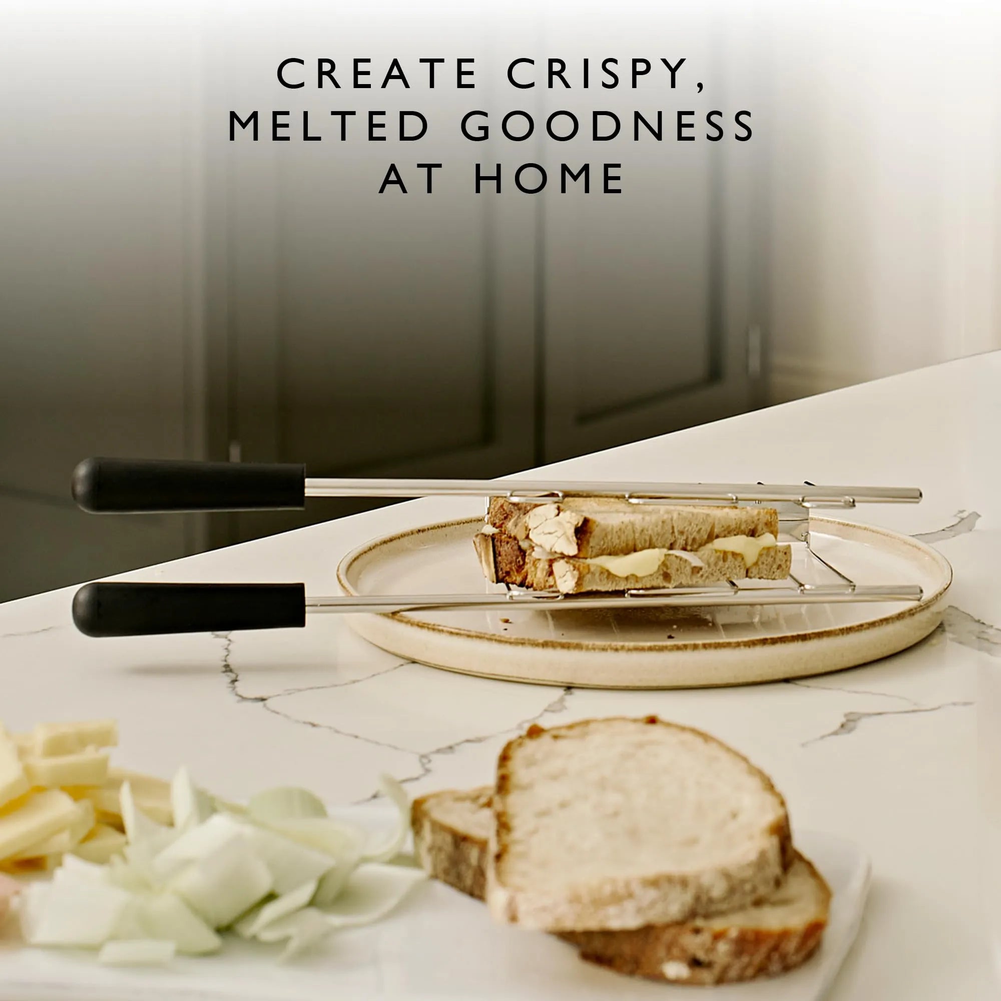 A close-up of a sandwich being prepared on a round plate using a metal sandwich cage with black handles, set on a white marble countertop with sliced bread, onions, and cheese nearby. Text at the top reads, 'CREATE CRISPY, MELTED GOODNESS AT HOME.