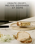 A close-up of a sandwich being prepared on a round plate using a metal sandwich cage with black handles, set on a white marble countertop with sliced bread, onions, and cheese nearby. Text at the top reads, 'CREATE CRISPY, MELTED GOODNESS AT HOME.