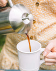 Person pouring coffee from a French press into a white mug outdoors.