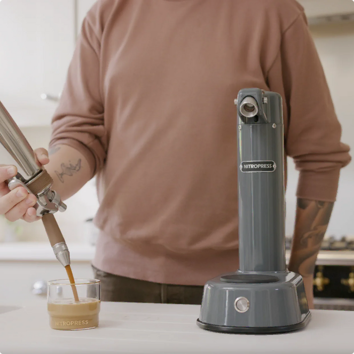 A person in a dusty rose long-sleeve shirt uses a metal pour-over spout to pour espresso into a small glass cup on a kitchen counter, with a gray Nitropress machine standing beside it.