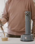 A person in a dusty rose long-sleeve shirt uses a metal pour-over spout to pour espresso into a small glass cup on a kitchen counter, with a gray Nitropress machine standing beside it.