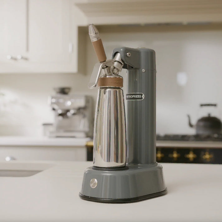 A countertop Electric Nitro Cold Brew Coffee & Cocktail Maker Machine with a matte gray body and base, a stainless steel bottle, and light wood accents, shown in a softly blurred kitchen setting.