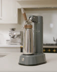 A countertop Electric Nitro Cold Brew Coffee & Cocktail Maker Machine with a matte gray body and base, a stainless steel bottle, and light wood accents, shown in a softly blurred kitchen setting.