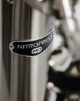 Close-up of a metallic NitroPress Pro coffee machine component with a black oval logo badge.