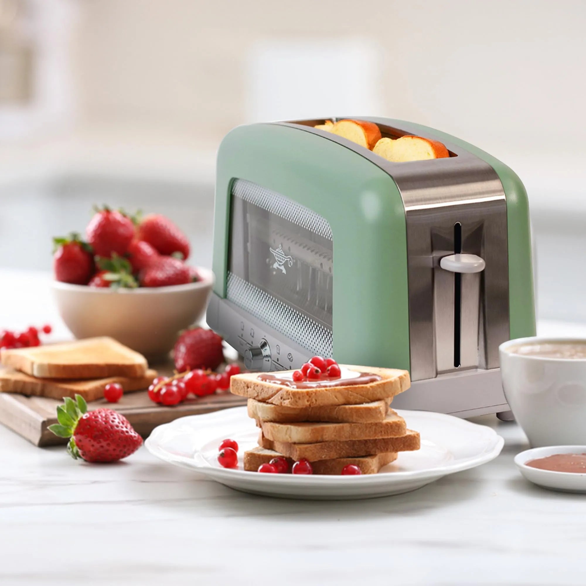 A retro-style two-slice toaster in sage green and stainless steel sits on a kitchen counter with toast, strawberries, and a cup nearby, suggesting a breakfast scene.